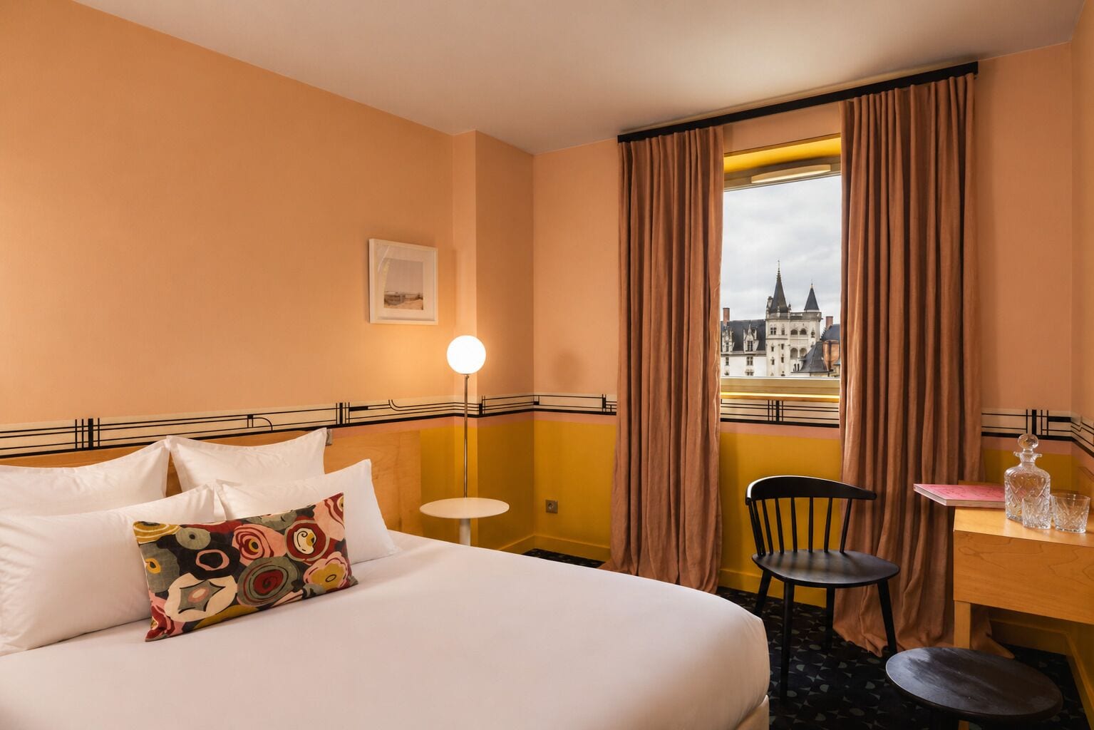 A hotel room with a bed, a desk, a chair, a small table, a lamp, and a window with curtains.