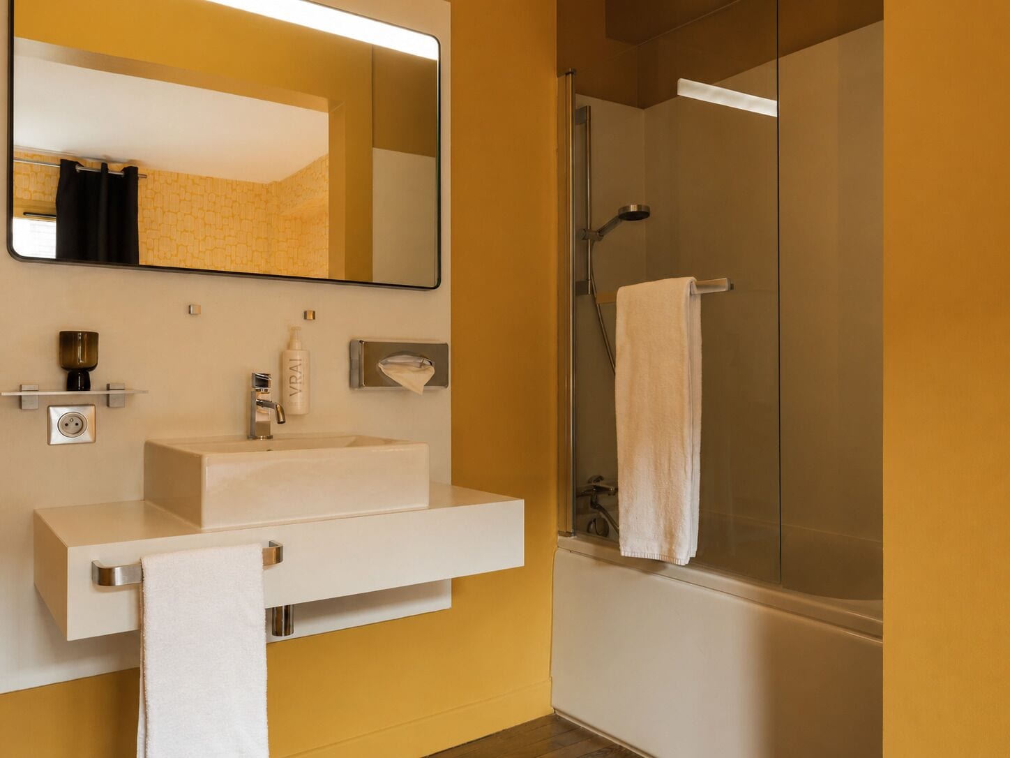 Un baño con lavamanos blanco, un espejo, toallero y zona de ducha con mampara de vidrio.
