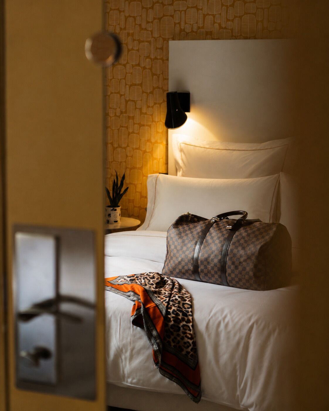 Habitación de hotel con una cama, una lámpara, una maleta y un objeto estampado de leopardo sobre la cama.