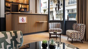 A modern hotel lobby with a reception desk, patterned chairs, a glass table, and a large window offering a view of the city. - Le 46 Hotel (Paris)