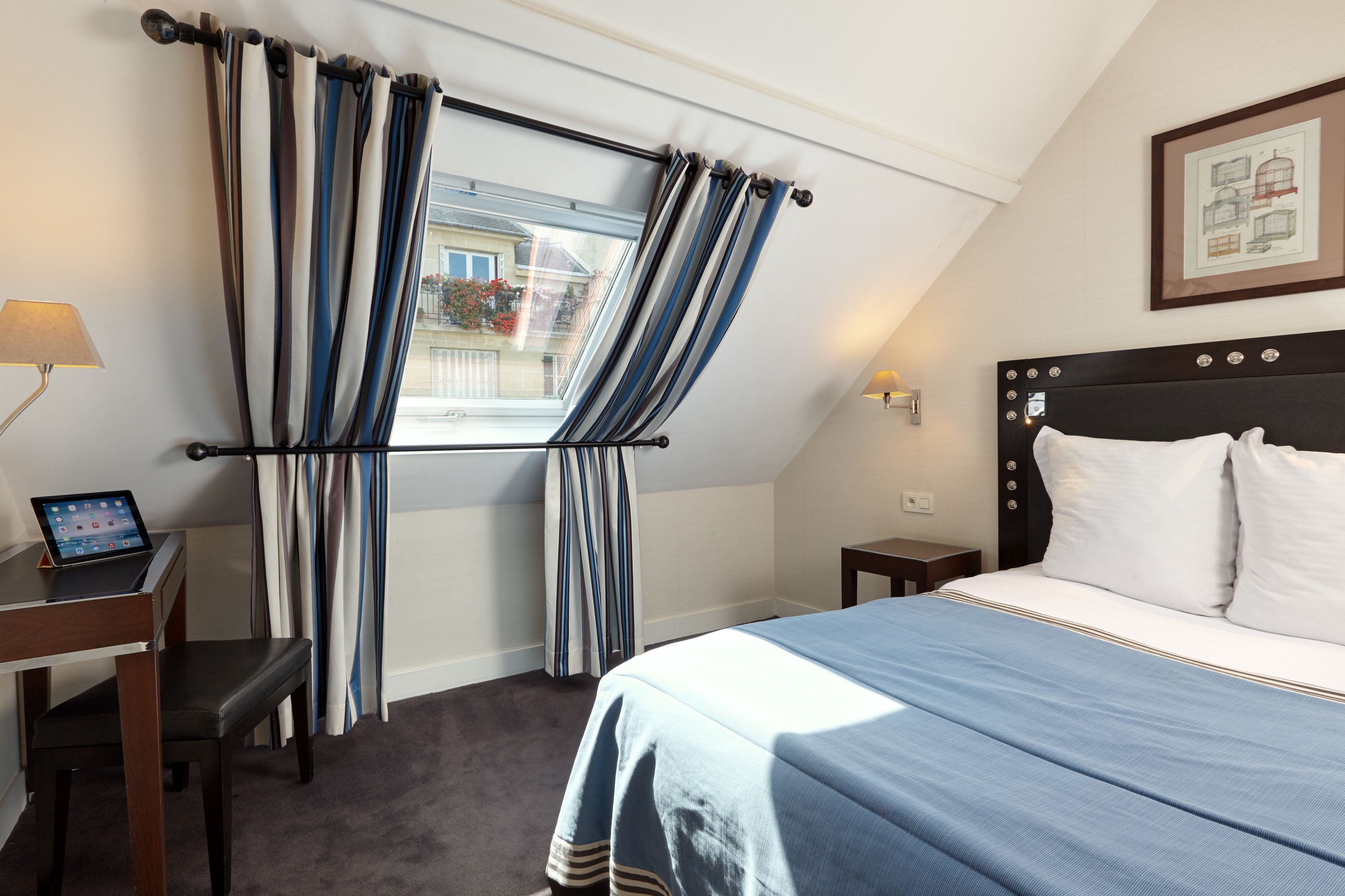 A hotel room with a bed, a desk with a laptop, a chair, a window with curtains, a bedside table, and a framed picture on the wall.