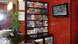 Zona de recepción con una estantería llena de guías de viaje, un televisor que muestra un noticiero y un refrigerador con bebidas.