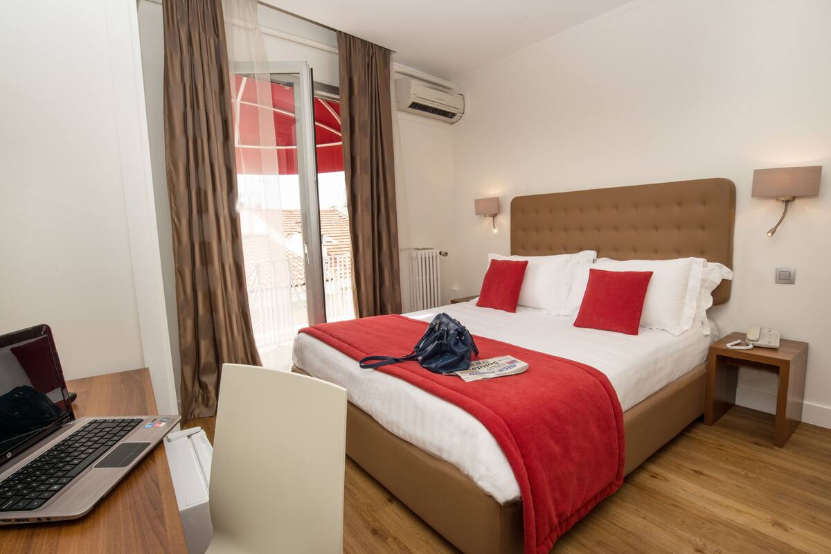 Ein Hotelzimmer mit Bett, Schreibtisch mit Laptop, Stuhl, Nachttisch mit Telefon und Fenster mit Vorhängen.