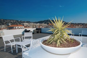 Una terraza en la azotea con una jardinera grande, sillas blancas y vistas a la ciudad y las colinas al fondo.