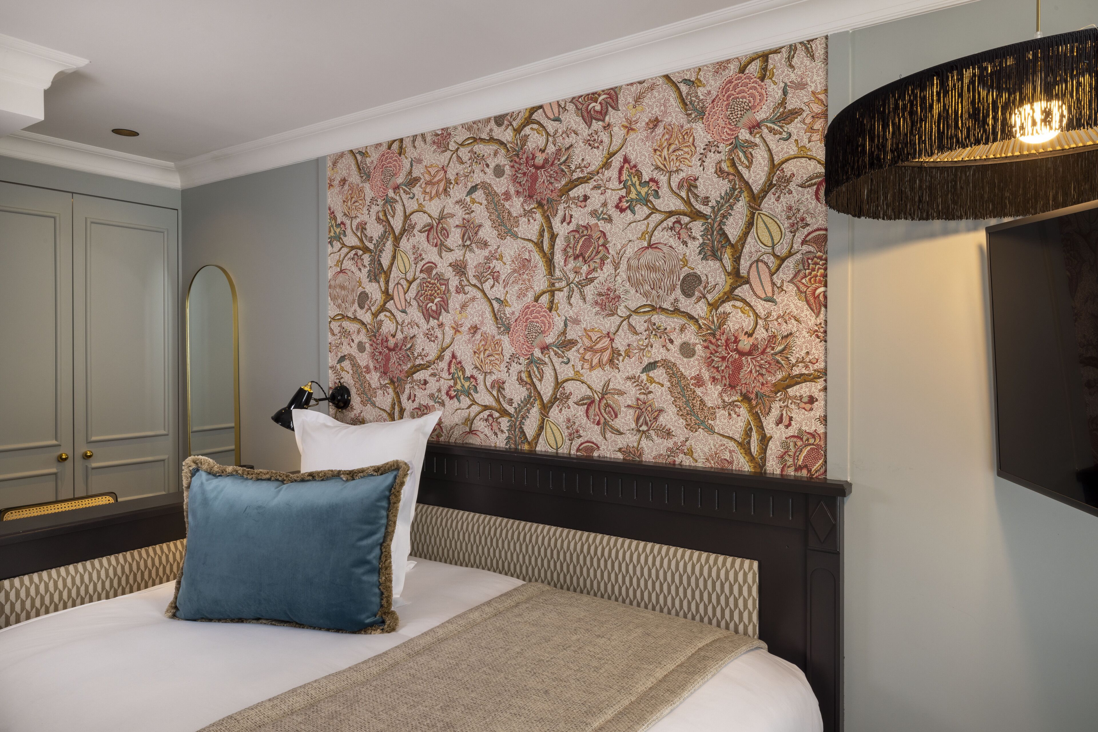 Una cama bien hecha con un cojín azul y un plaid color beige, una pared con estampado floral y un cabecero de madera oscura.