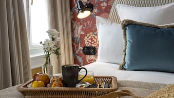 Un plateau de petit-déjeuner avec des fruits, une tasse et un verre de jus d’orange, posé sur un lit dont le papier peint est à motifs floraux, avec une lampe de chevet et un oreiller bleu.