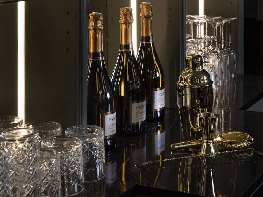 Una zona de bar con varias botellas de vino, copas de vino y un decantador dorado sobre una superficie reflectante.