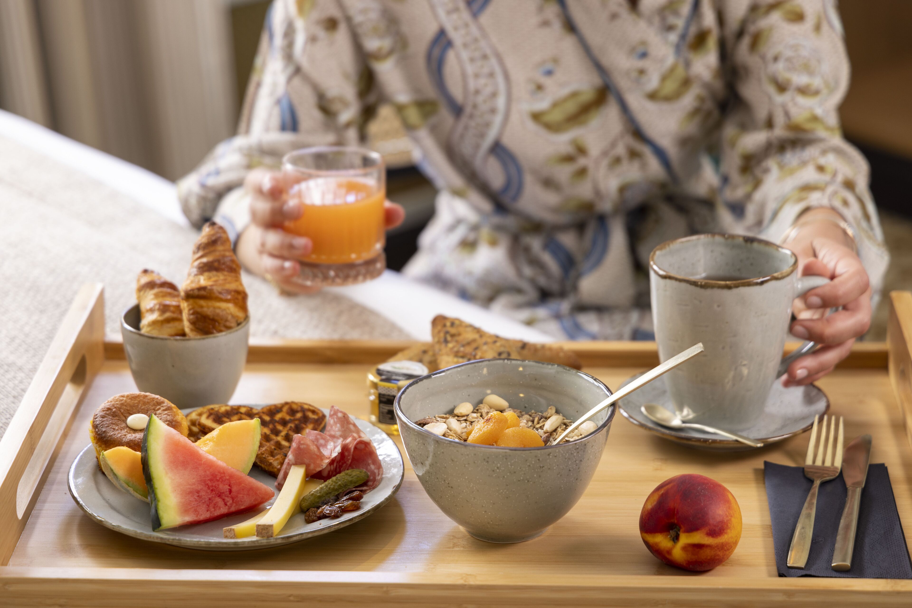 Een ontbijttafel met croissants, wafels, een kom ontbijtgranen, een glas sinaasappelsap en een bord met fruit en vlees.