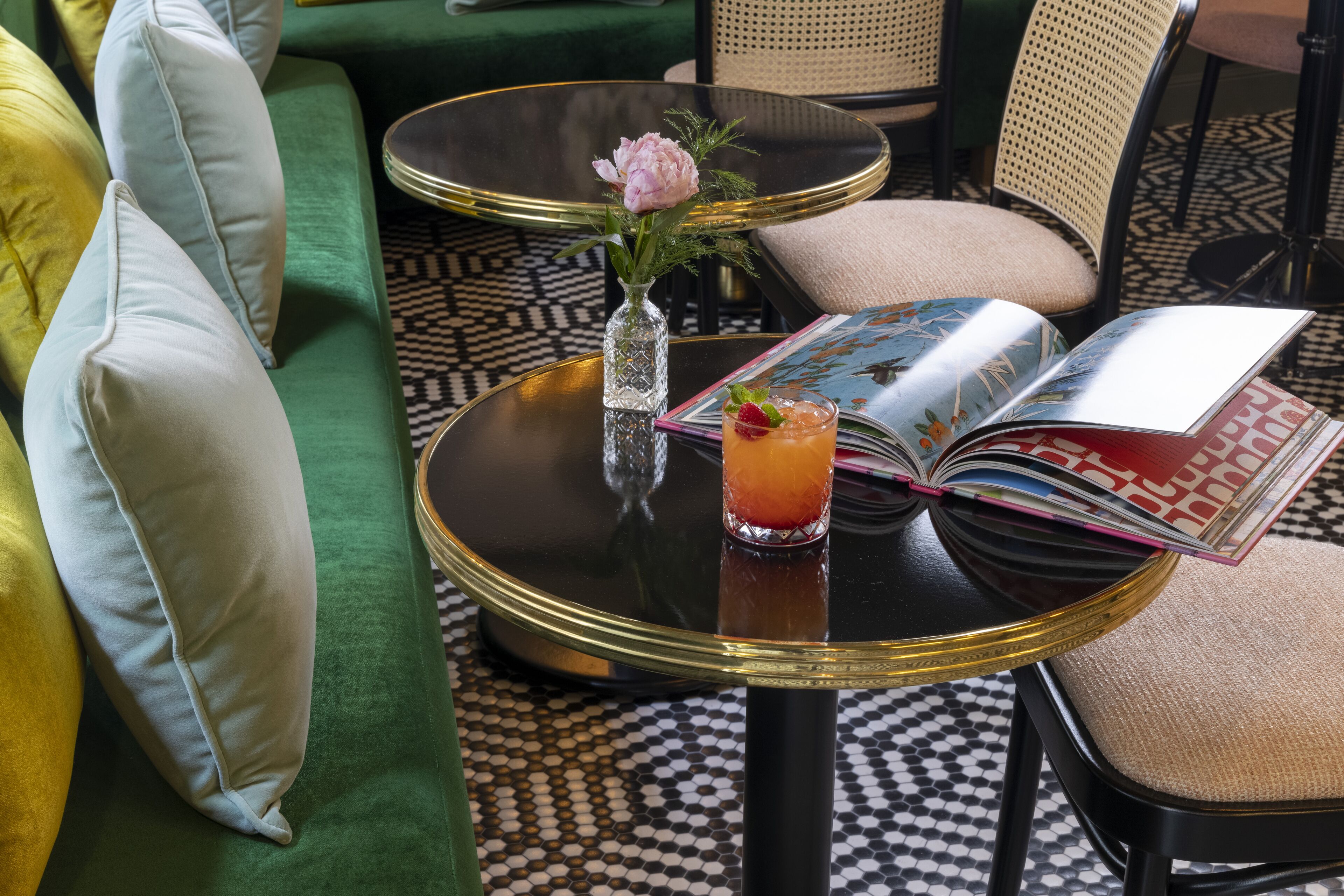A green sofa with light-colored pillows, a small round table with a glass vase and a drink, and an open magazine.