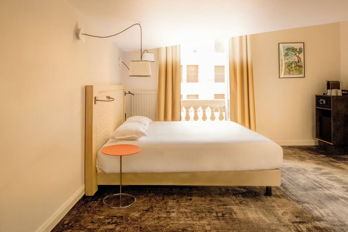Ein Hotelzimmer mit einem Bett, einem runden orangefarbenen Beistelltisch, einer Wandlampe, einer Heizung, einem Fenster mit Vorhängen und einem gerahmten Bild an der Wand.