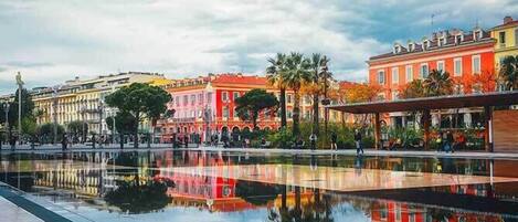 A plaza with a reflective water surface, colorful buildings, and palm trees.