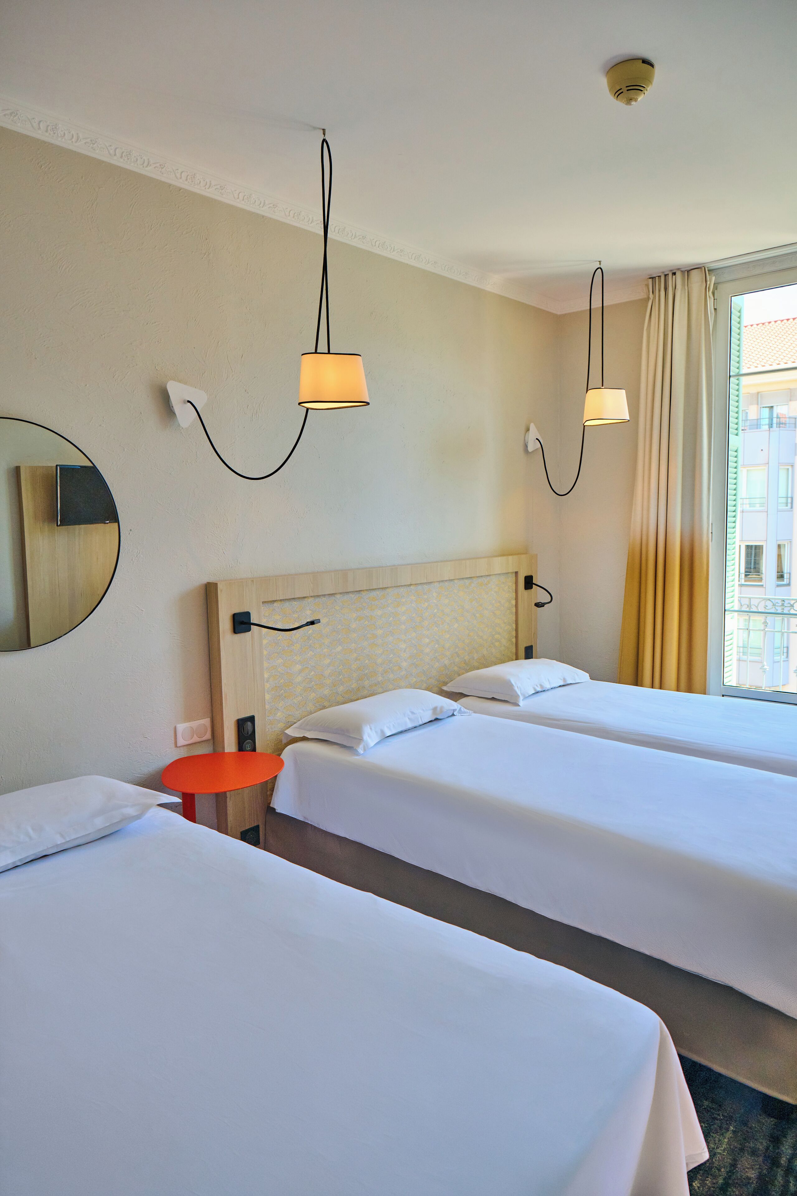 A hotel room with two beds, a round mirror, a small orange table, and a window with curtains.
