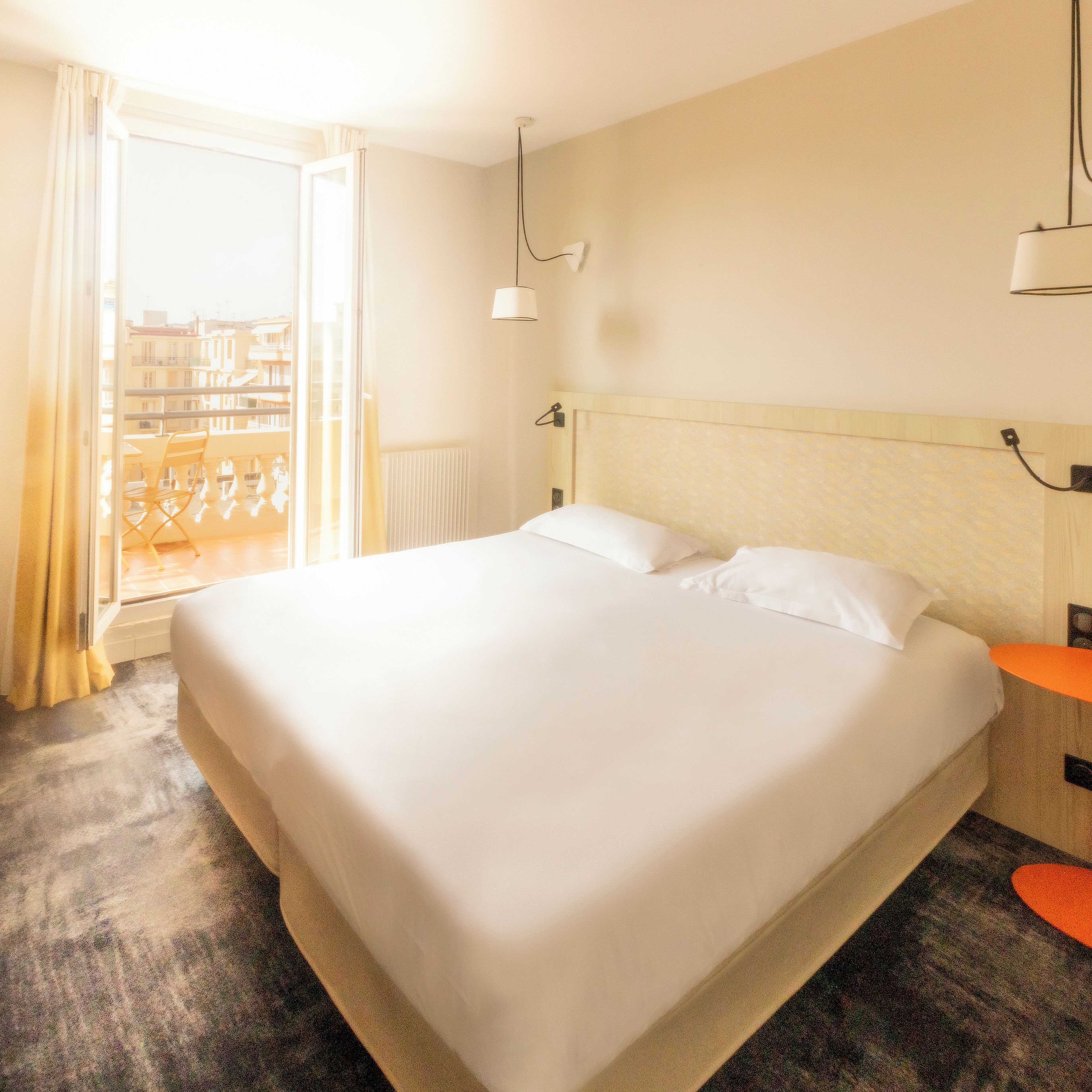 A hotel room with a large bed, a window with curtains, a radiator, and a small table with a lamp.