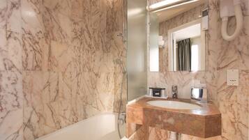 A bathroom with a marble wall, a white bathtub, a sink with a marble countertop, a glass shower enclosure, and a mirror with a hairdryer mounted on the wall.