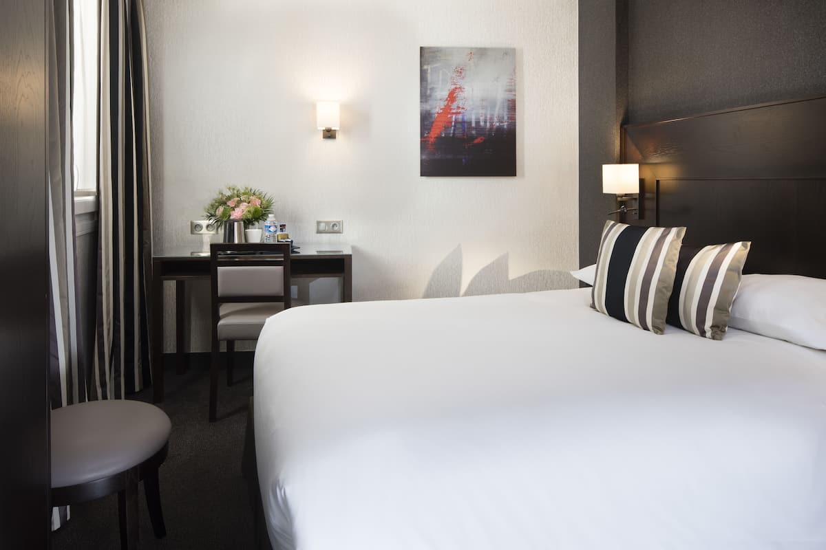 Une chambre d’hôtel avec un grand lit, un bureau avec une chaise, une petite table avec un vase de fleurs et un tableau au mur.