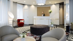 A modern hotel lobby with a reception desk, a red armchair, a round ottoman, and two grey armchairs. - Hôtel Pastel Paris (Paris)