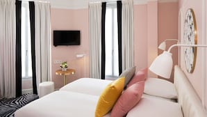 A hotel room with a bed, pillows, a bedside table, a lamp, a TV, and a window with curtains. - Hôtel Pastel Paris (Paris)