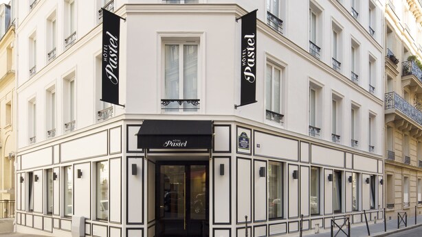 A corner building with a white facade, multiple windows, and balconies. There are two black banners with the text "HOTEL Pastel" hanging from the building. - Hôtel Pastel Paris (Paris)