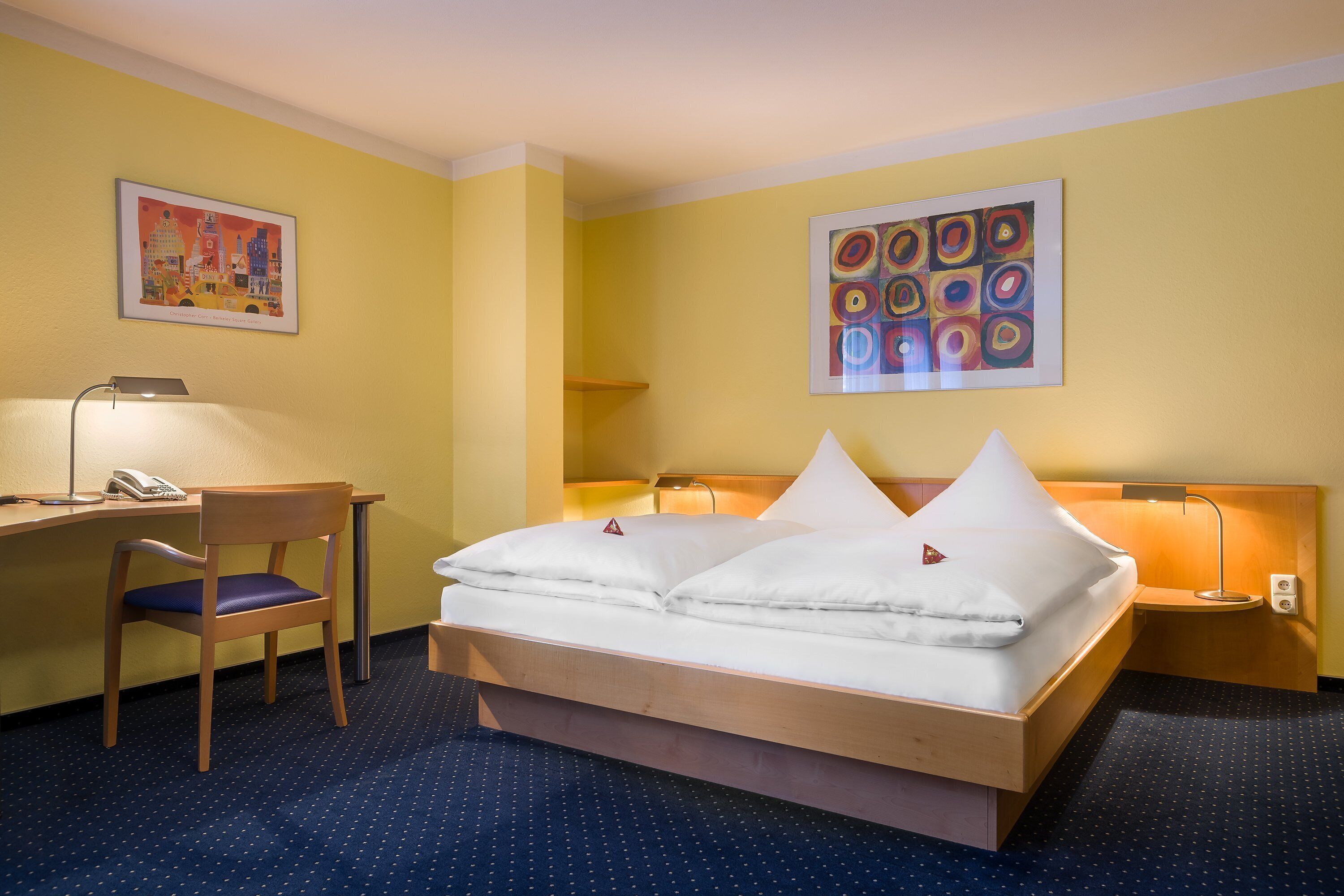 A hotel room with a bed, desk, chair, and two wall-mounted lamps.