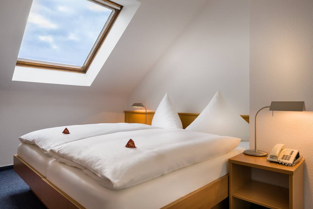 Una habitación de hotel con tragaluz, una cama con ropa blanca, una lámpara de noche y un teléfono sobre una mesita de madera.