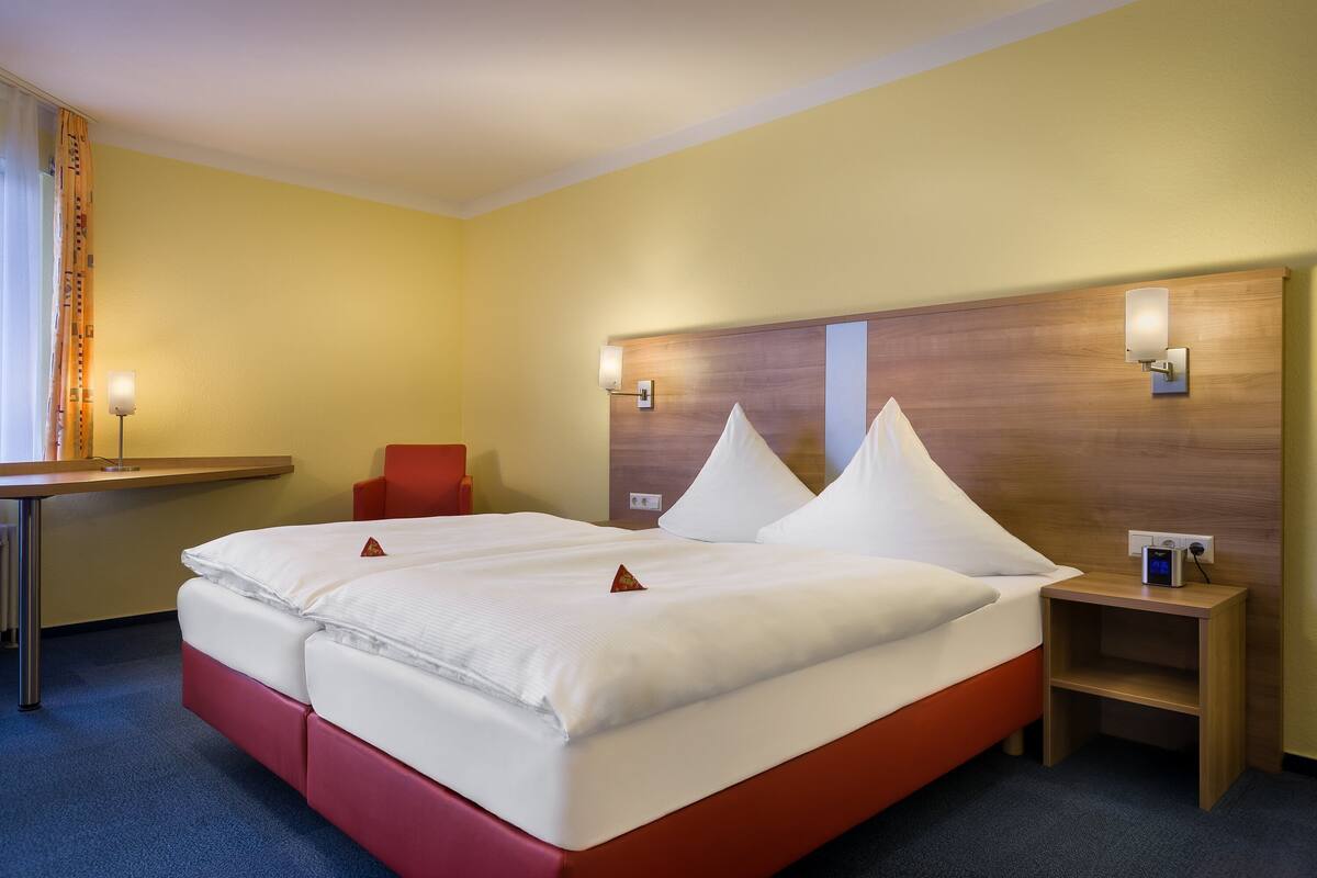 Habitación de hotel con cama doble, cabecero de madera, mesita de noche con lámpara y una silla roja.