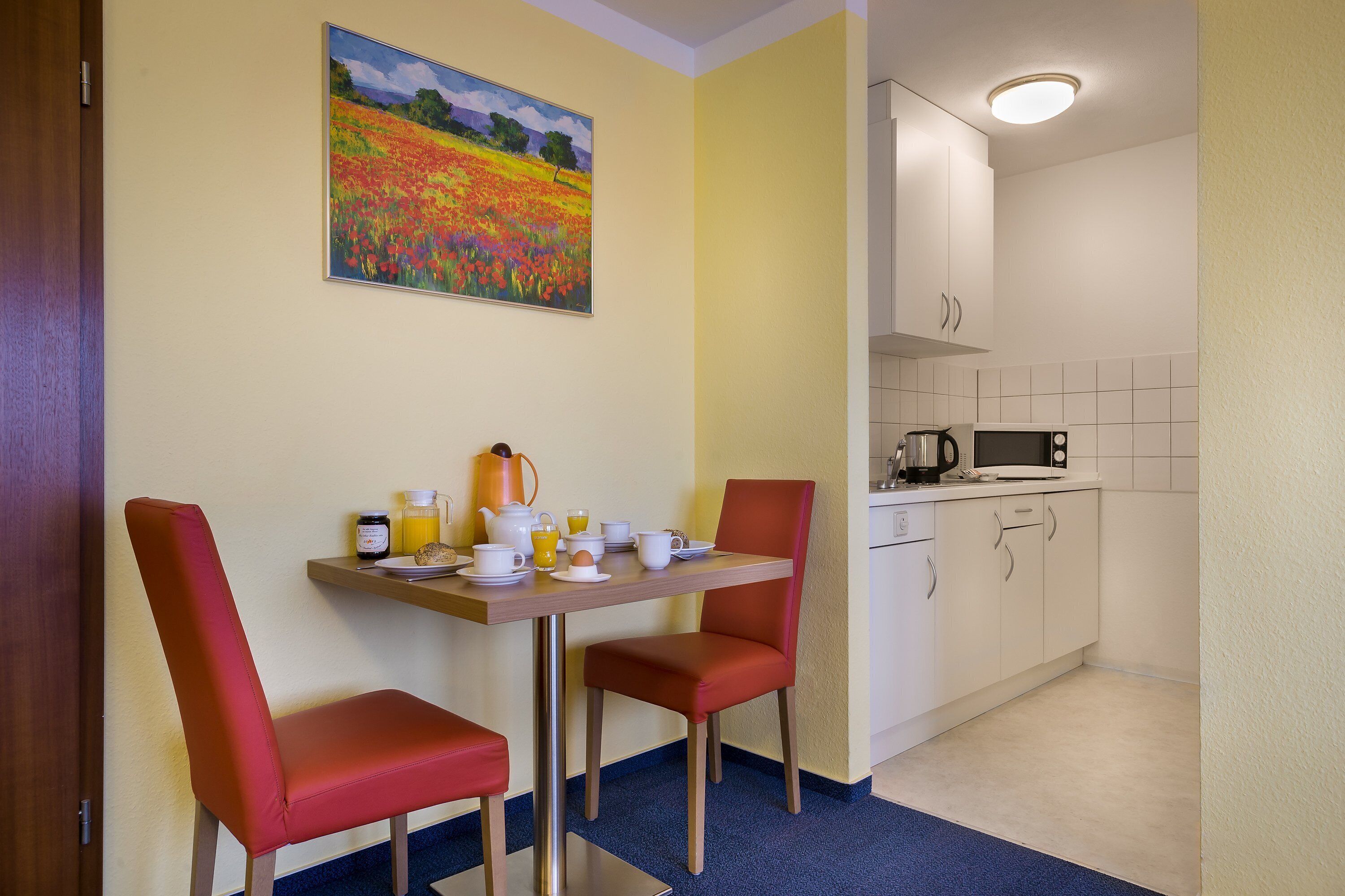 A compact room with a dining table set for breakfast, two red chairs, a painting on the wall, and a kitchenette area.