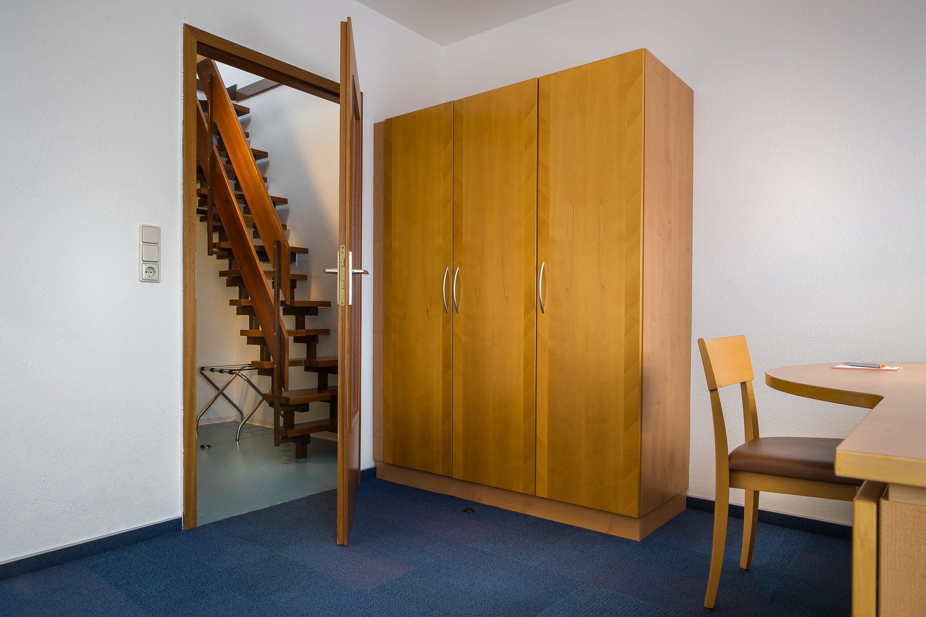 A room with a wooden wardrobe, a dining table with a chair, and a staircase.