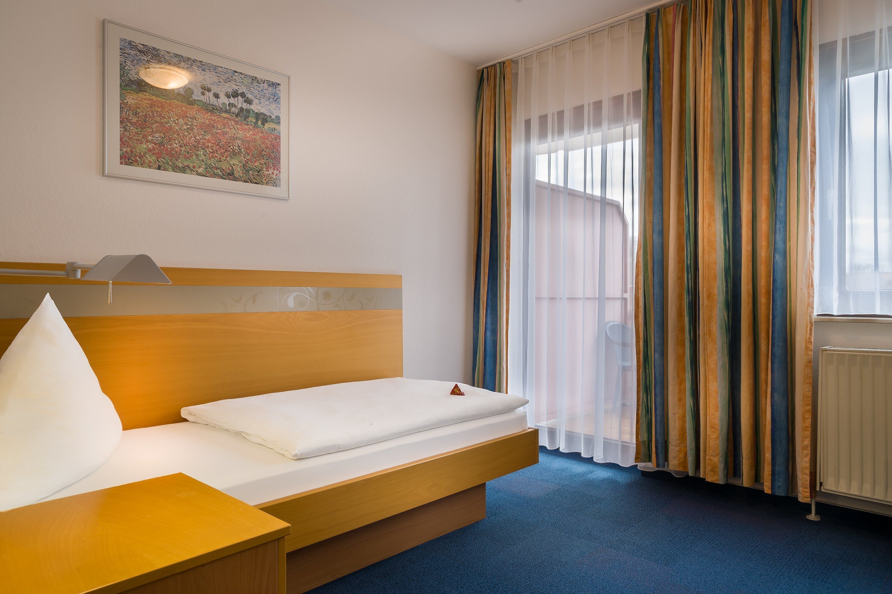 A hotel room with a bed, a bedside table, a lamp, a window with curtains, and a painting on the wall.