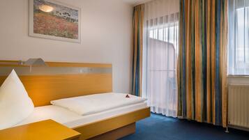 A hotel room with a bed, a bedside table, a lamp, a window with curtains, and a painting on the wall.