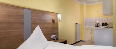 A hotel room with a wooden headboard, a white pillow, a bedside table with a lamp, and a kitchenette in the background.