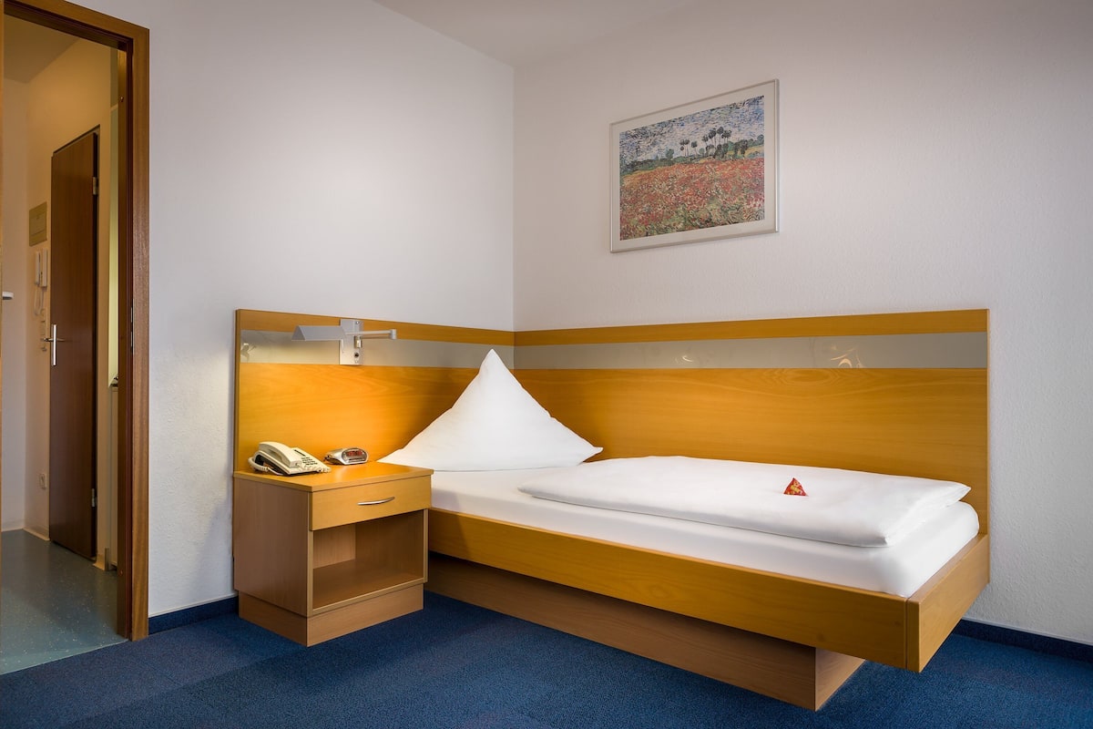 Una habitación de hotel con una cama de madera, una mesita de noche con un teléfono, un cajón pequeño y una almohada blanca, además de un cuadro en la pared.