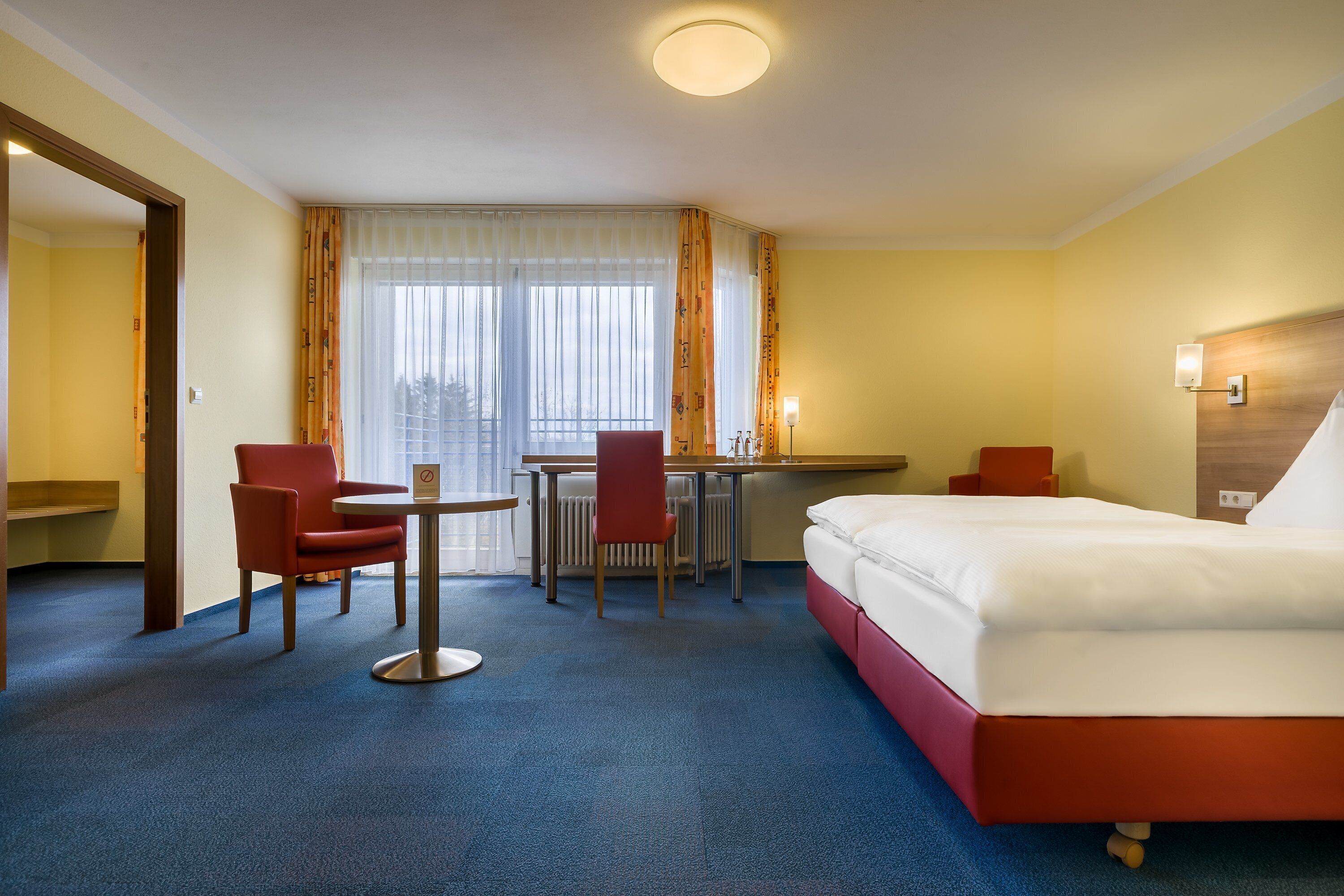 Una habitación de hotel con una cama, dos sillas rojas, una mesita y una ventana con cortinas.