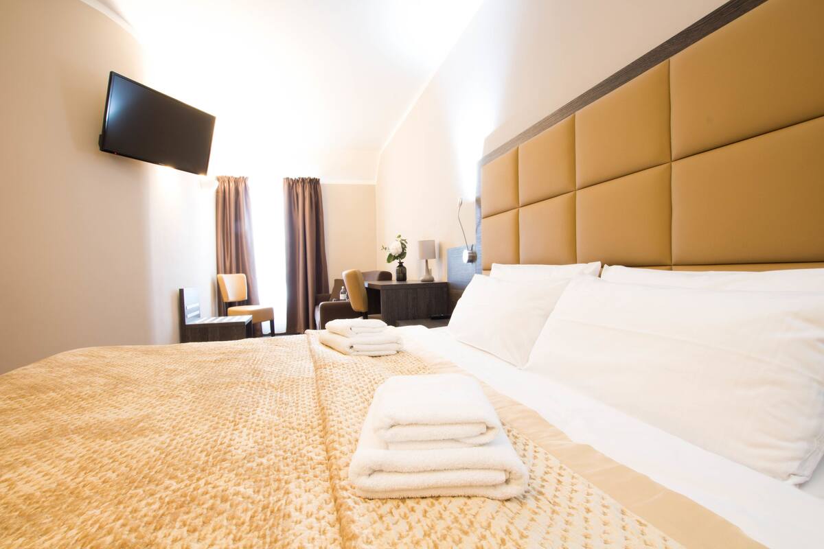Habitación de hotel con una cama grande, un televisor de pantalla plana montado en la pared, un escritorio con una silla y una mesita de noche con una lámpara.