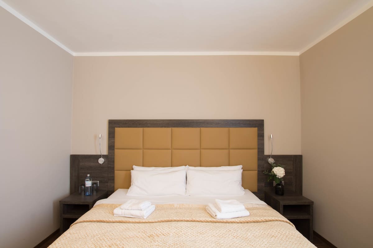 Una cama bien hecha con ropa y toallas blancas, cabecera de madera y una mesita de noche con una botella de agua y un florero.