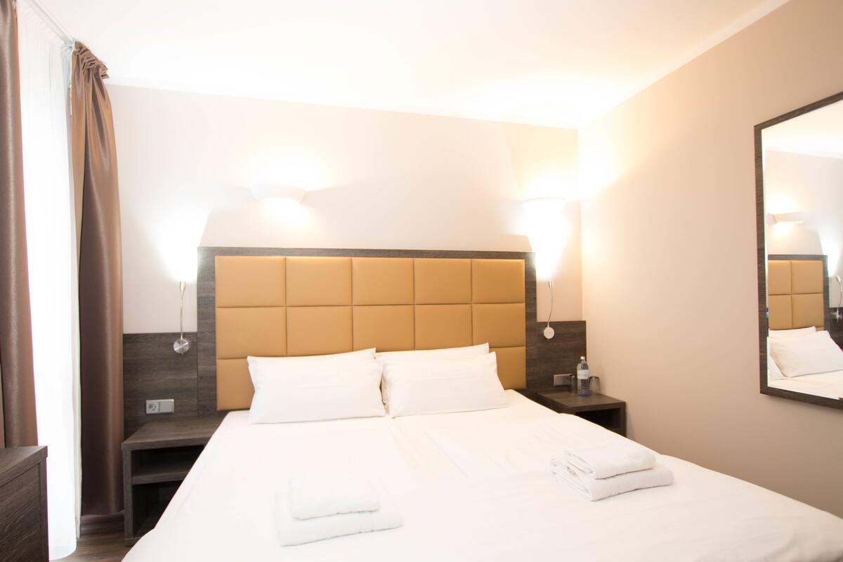 Habitación de hotel con una cama grande, dos mesitas de noche, una lámpara de noche, un espejo y una ventana con cortinas.