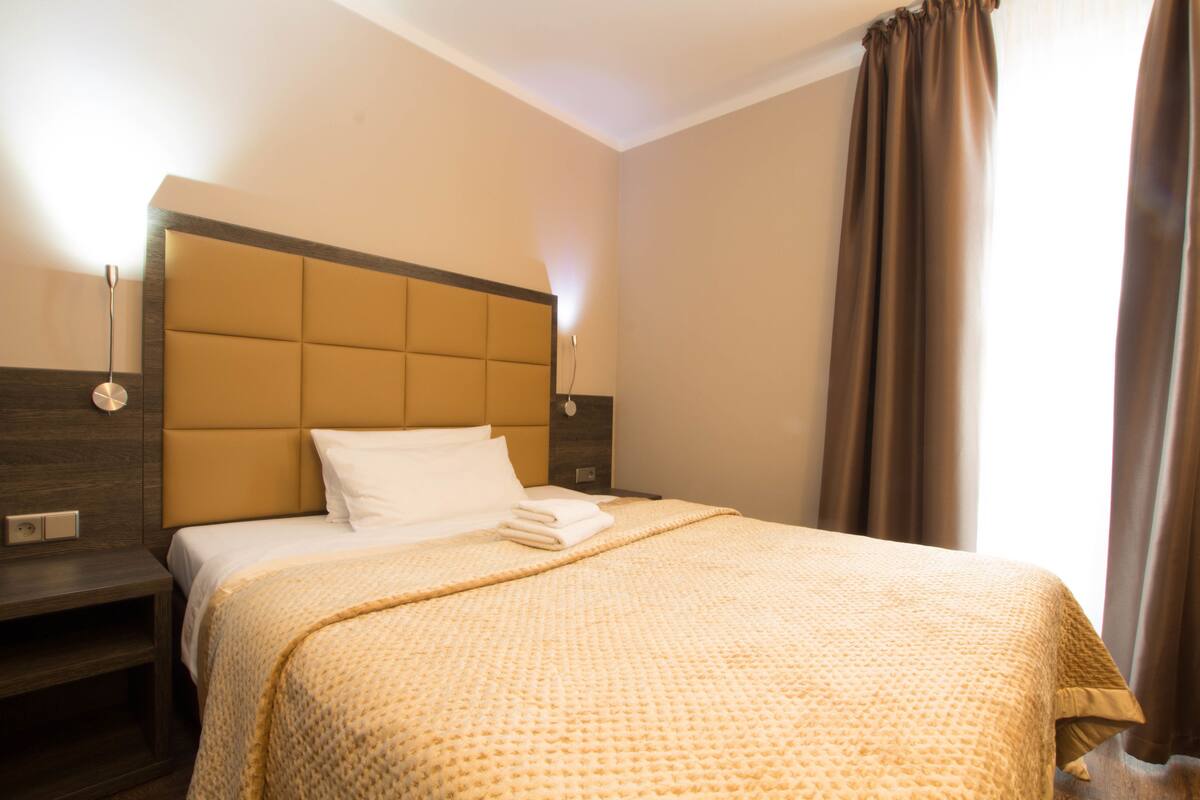 Una habitación de hotel con una cama grande, un cabecero, mesitas de noche y una ventana con cortinas.