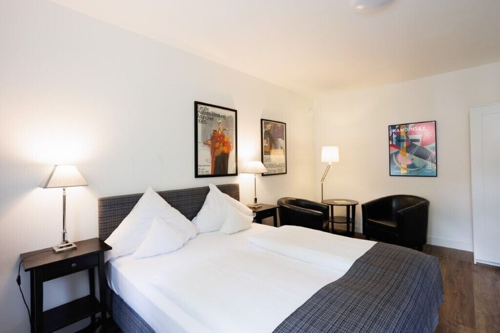 Una camera d'albergo con un letto, due comodini con lampade, una sedia e opere d'arte alle pareti.