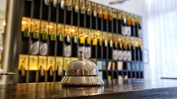Un espace bar doté d’un comptoir en bois poli et d’une clochette argentée pour signaler le service. Derrière le comptoir, on trouve des étagères remplies de différentes bouteilles et une horloge murale.