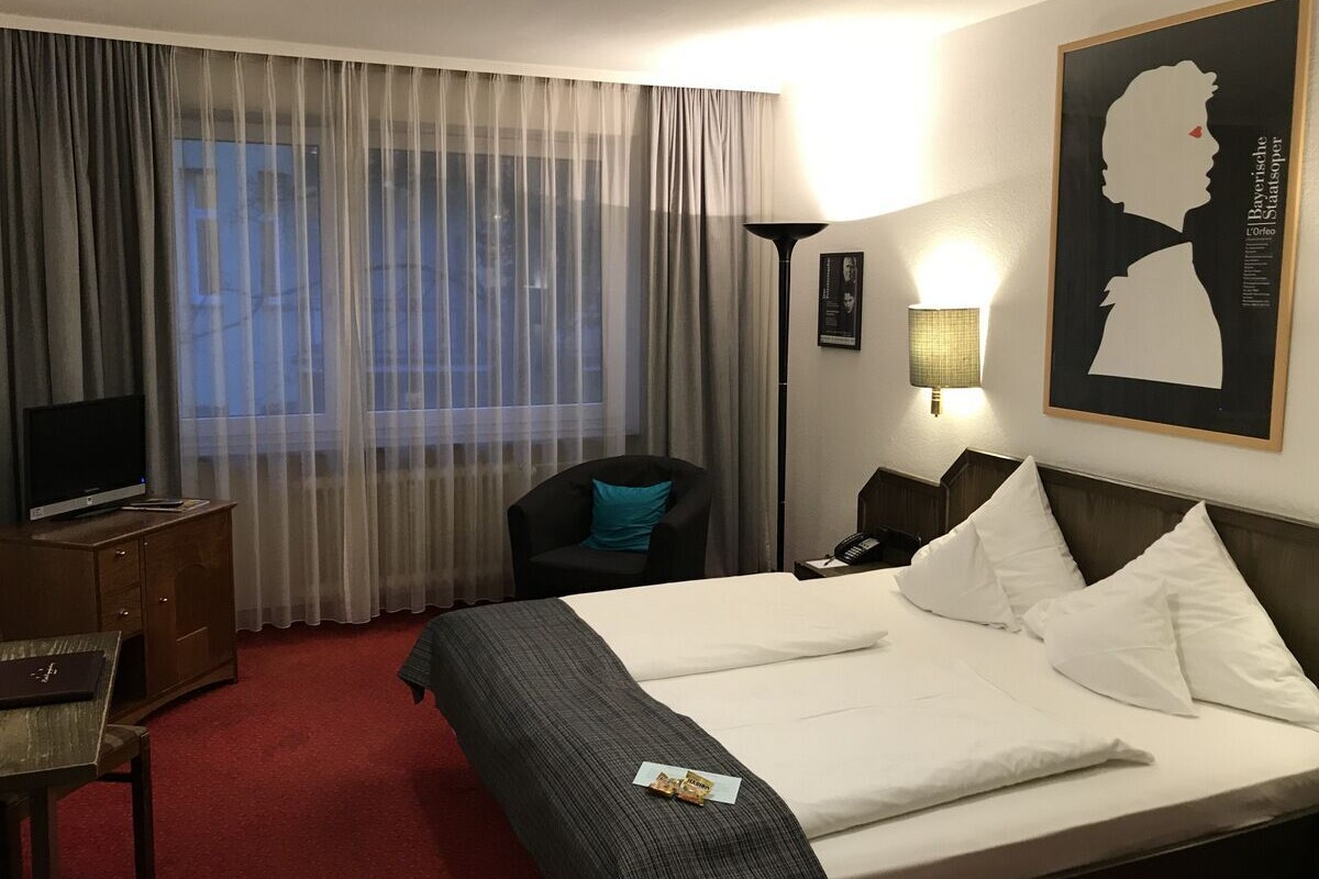 A hotel room with a large bed, a desk with a TV, a chair, and a window with sheer curtains.