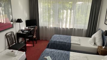 Une chambre d’hôtel avec deux lits, un bureau équipé d’un ordinateur et d’une lampe, et une grande fenêtre avec des rideaux.