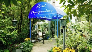 Une tente bleue sur laquelle on peut lire « Hôtel Nymphengurg », entourée de verdure et avec un banc  à proximité.