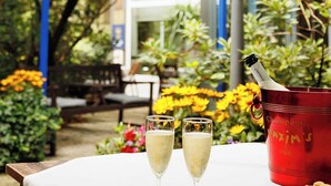 Une table avec deux coupes à champagne et un seau rouge portant l’inscription « Champagne Saxin’s », le tout installé dans un jardin agrémenté d’un parasol bleu et d’une pancarte indiquant « Hôtel Nymphenburg ».