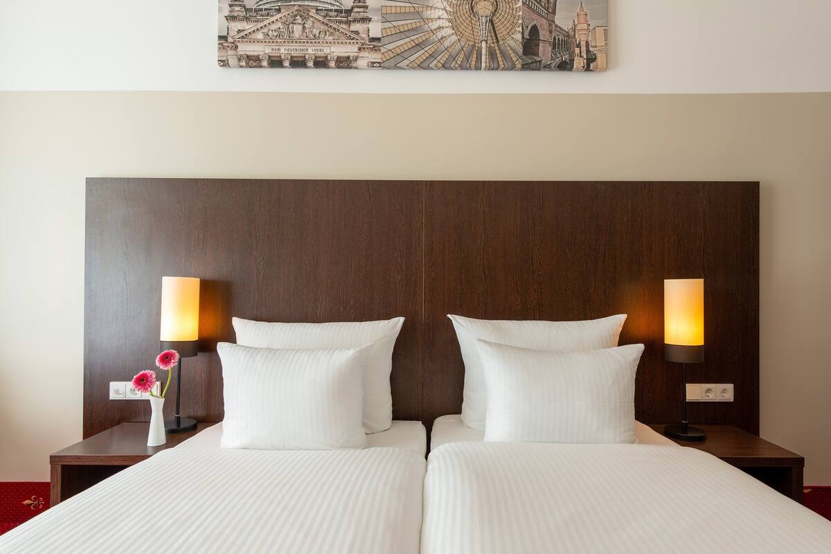Uma cama de casal com lençóis brancos, duas luminárias de cabeceira e um vaso com flores sobre um criado-mudo de madeira.