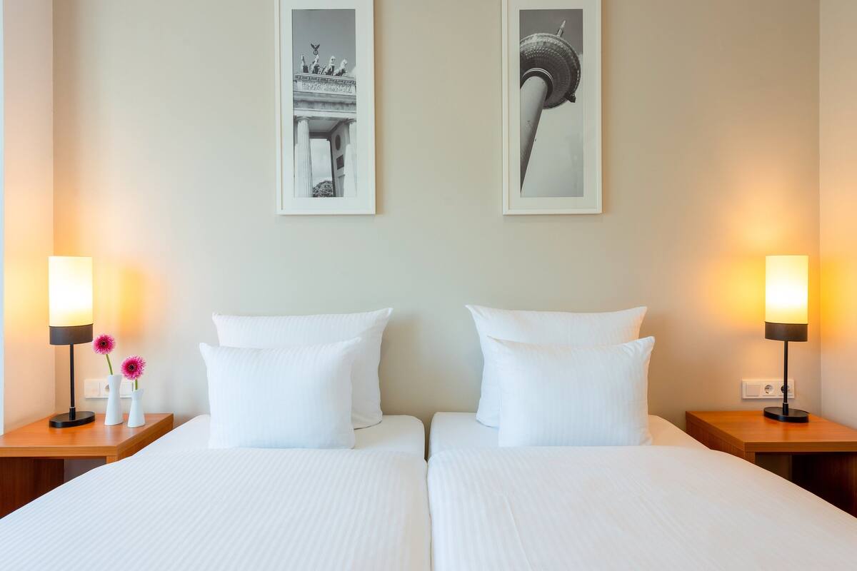 Quarto de hotel com duas camas, roupa de cama branca e mesas de cabeceira com luminárias e vasos.