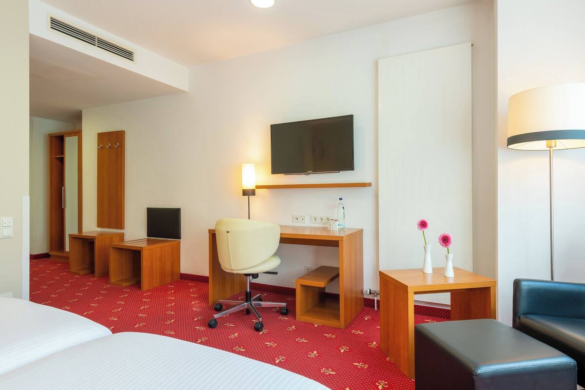 A hotel room with a bed, a desk with a chair, a television, a lamp, and a small table with flowers.