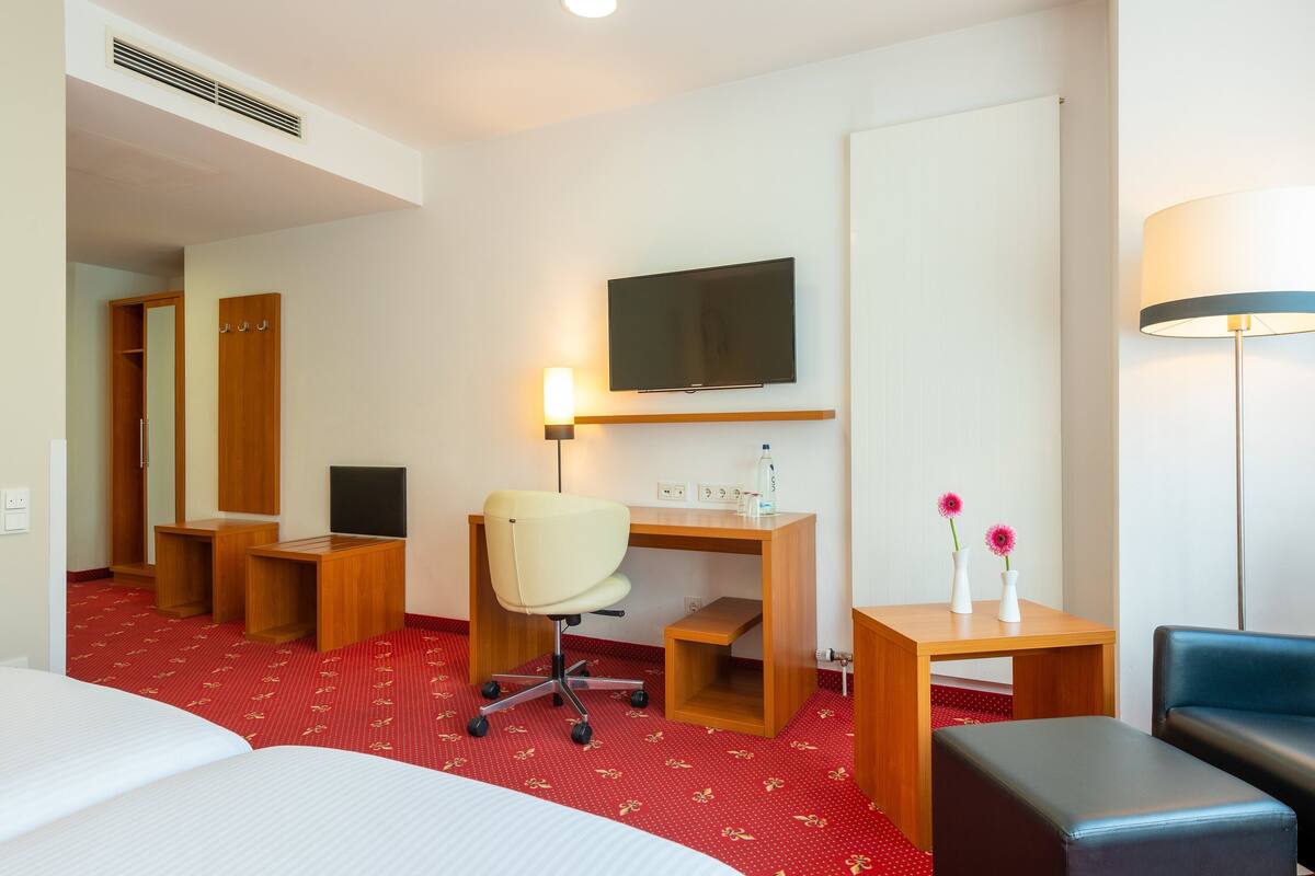 Quarto de hotel com uma cama, uma escrivaninha com cadeira, televisão, abajur e uma pequena mesa com flores.