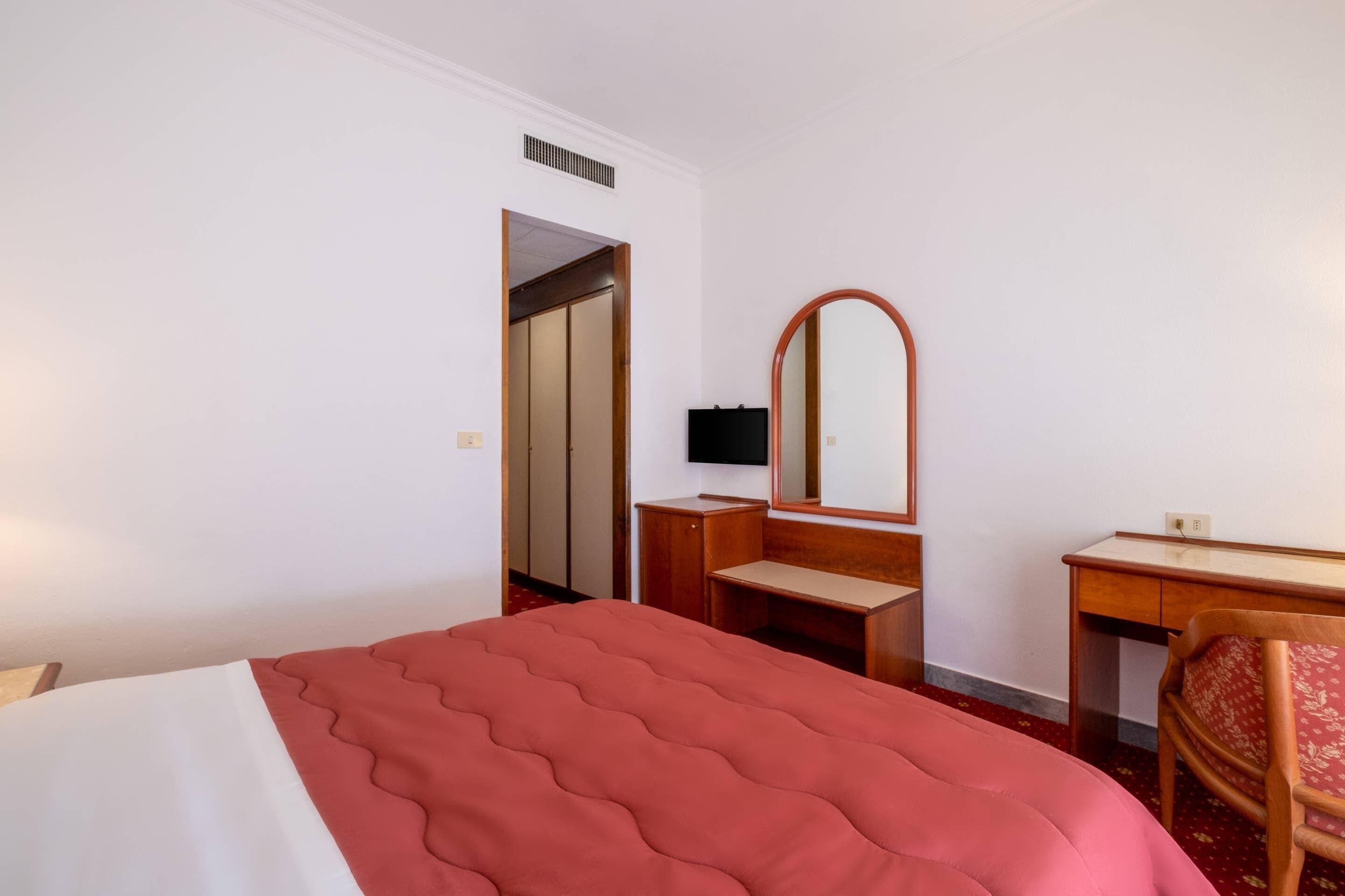 A hotel room with a bed, a desk, a chair, a mirror, and a wardrobe.