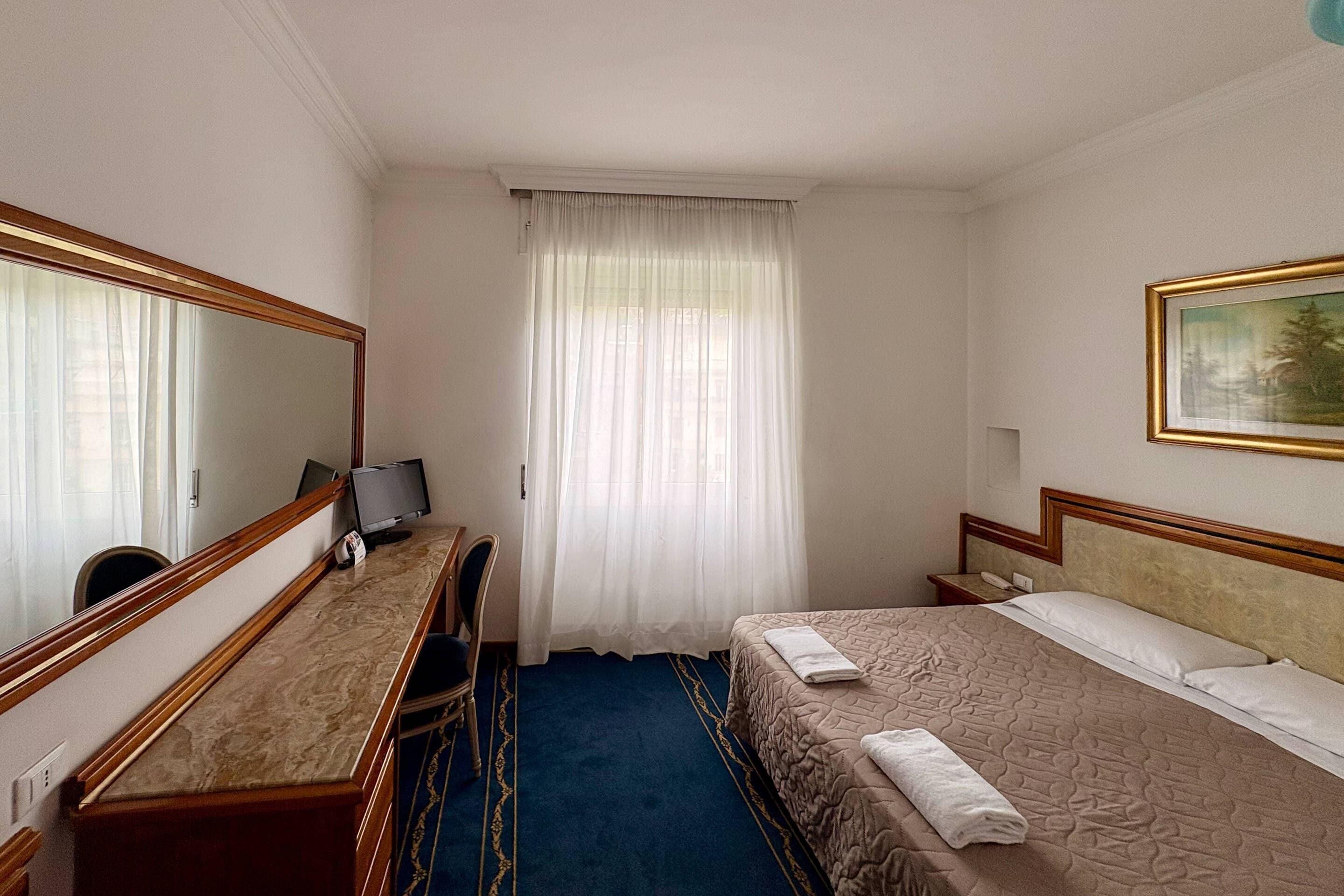 A hotel room with a bed, a desk with a chair, a television, and a large mirror.