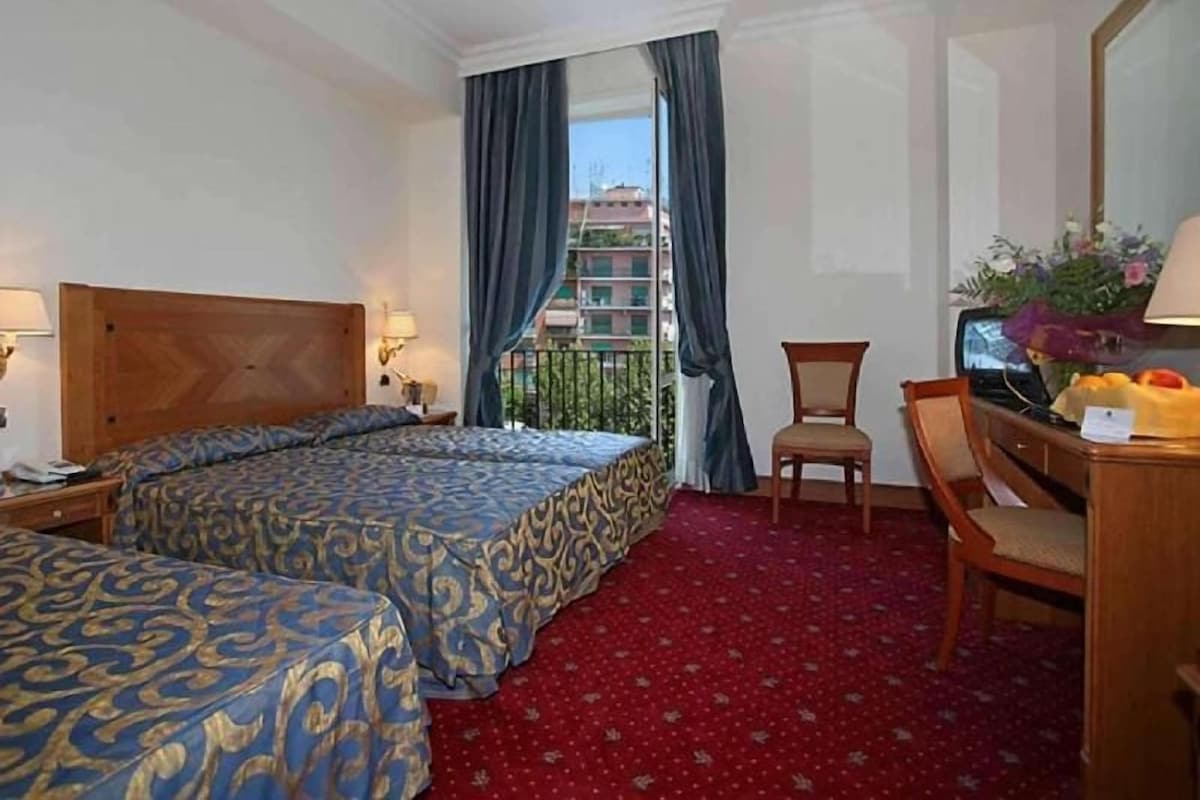 A hotel room with two beds, a desk with a chair, a TV, and a balcony with curtains.