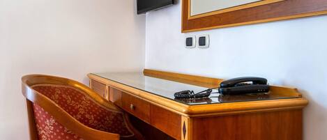 A wooden desk with a telephone, a chair with a floral pattern, a television mounted on the wall, and two electrical outlets.